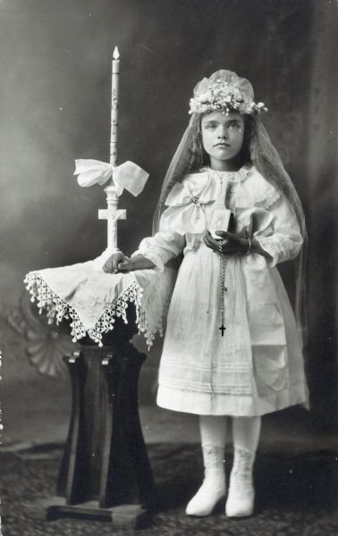 Photographic postcard of a studio portrait of a girl on the occasion of her First Communion. She is dressed in a white dress and flowered tiara and holds a rosary and catechism in her left hand. She is posed in front of a painted backdrop.
