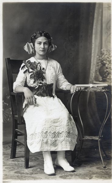 Photographic postcard of a seated studio portrait of Rose Worringer on the occasion of her First Communion. She has a decorative bouquet of flowers pinned to her chest and is wearing a white dress and a large bow in her hair. A rolled paper certificate rests on the table by her left hand. She is sitting in a chair and is posing in front of a painted backdrop.