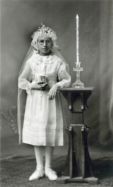 Photographic postcard of a full-length studio portrait of an unidentified girl on the occasion of her First Communion. She is wearing gloves, a white dress, flowered headpiece with veil, and is holding a rosary and catechism in her right hand. She is standing in front of a painted backdrop and is resting her left elbow on a table which has a glass candle holder in the shape of a cross, with a tall decorative candle that appears to be lit.