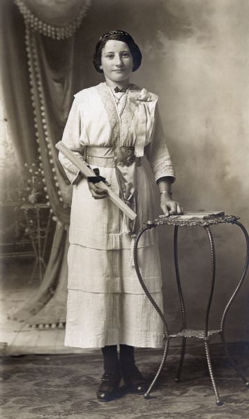 Photographic postcard of a full-length studio portrait of Carolyn Zander Ziegler on the occasion of her graduation. She is standing and is wearing a white dress and is holding a rolled diploma in her right hand. She is resting her left hand on a table and is posing in front of a painted backdrop.