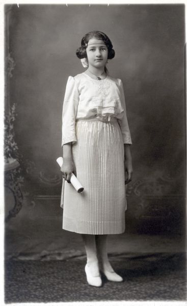 Photographic postcard of a full-length studio portrait of Viola Festge Hering on the occasion of her graduation. She is standing and is wearing a pleated white skirt and white shoe. She has a hairnet over her forehead and has a large bow in her hair. She is holding a rolled diploma in her right hand and is posing in front of a painted backdrop.