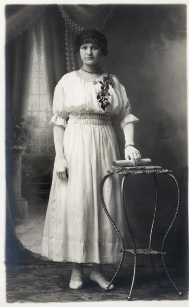 Photographic postcard of a full-length studio portrait of a young woman on the occasion of her graduation. She wears a white dress with a corsage pinned to her bodice and holds a rolled diploma in her left hand. She is posed in front of a painted backdrop.