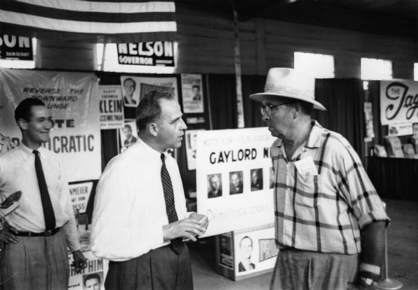 Nelson talks with a gentleman at a campaign booth, at the State Fair, during his run for Governor. In the background there are several campaign signs endorsing Democratic candidates Proxmire and Nelson.