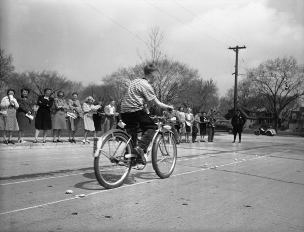 Side view of a Marquette School student being tested for riding skills in front of the school at 510 South Thornton Avenue. The testing is along Spaight Street. A group of adults are lined up on the sidewalk watching.