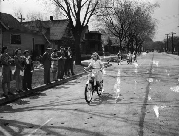 Marquette School girl student being tested for riding skills in front of the school at 510 South Thornton Avenue. The testing is along Spaight Street. A group of adults are lined up on the sidewalk watching.