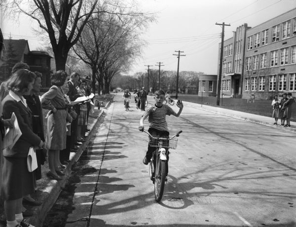 Marquette School student, signaling a left turn, being tested for riding skills in front of the school at 510 South Thornton Avenue. The testing is along Spaight Street. Other students on bicycles wait in the background.