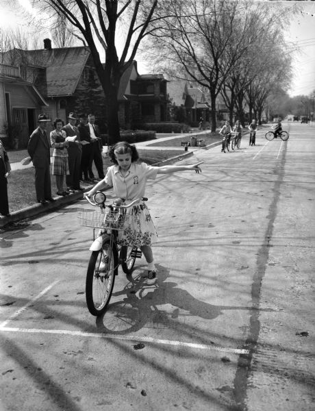 Marquette School student, a girl in a flowered skirt, being tested for riding skills in front of the school at 510 South Thornton Avenue. The testing is along Spaight Street. Other students on bicycles wait in the background.