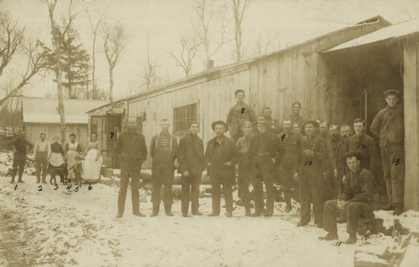 Shingle mill workers posing outside the cook shanty while the mill was shut down for repairs. They are, left to right as numbered on the photograph: 1. John Marsten 2. Peter Becker 3. Mrs. John Colombo 4. Mrs. Peter Becker 5. Milton Ritter 6. Mrs. John Ritter 7. the photographer's wife 8. Christian Hanson 9. Jas Clark 10. Barney Albee 11. Frank Baker 12. Red Merrison 13. Arnold Becken. The other men are unidentified.