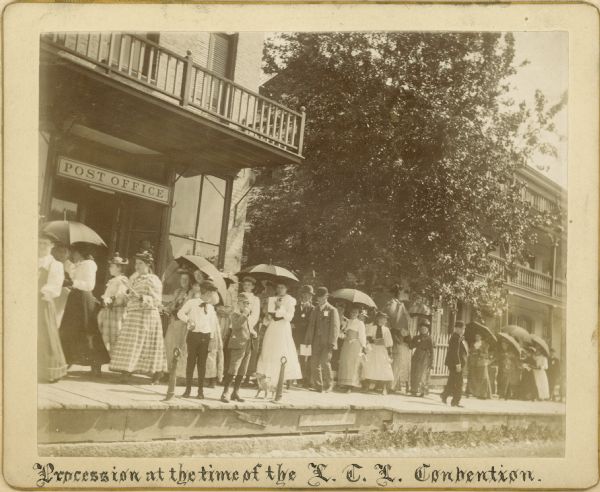 Parade of men, women, and young people attending the second convention of the Wisconsin Loyal Temperance Union at Delavan.  The LTL was a branch of the Women's Christian Temperance Union that focused on the temperance education of children. They are walking on the wooden sidewalk near the post office.  Many people are carrying umbrellas to protect themselves from the sun.