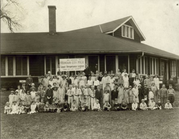 At a convention of the Fond du Lac County chapters of the Loyal Temperance League, some of the children present for a group picture hold up a sign that says, "We Drink Fountain City Milk."  The picture is taken in front of the community building.