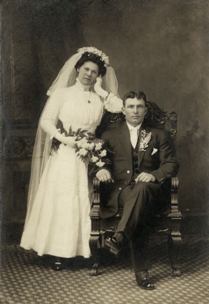 Studio portrait in front of a painted backdrop of Rose and Louis Meyer. Rose Swetlik (1892-1968) married Louis Meyer (1887-1949), a farmer, on February 28, 1911 in Kossuth Township (Manitowoc Co.). The wedding party then drove to Manitowoc to have their photographs taken. Bride is standing, groom is seated, both are wearing formal wedding attire.