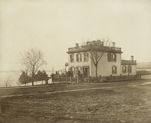 Early view of the house built by Madison's first mayor, Jairus C. Fairchild, at 302 South Wisconsin Avenue (renamed Monona Avenue in 1877) at West Wilson St. near Lake Monona. There is a woman standing near the front door. The house was constructed in 1850 and later was substantially altered with additions. After the Civil War, the house became the home of Fairchild's son, Lucius, Civil War hero, Wisconsin governor (1866-72) and foreign diplomat. During his six years as governor, the Fairchild House was the state's executive residence, as no such property was then owned by the state. The residence was therefore a focus of Madison's political and social life during the last half of the 19th century.