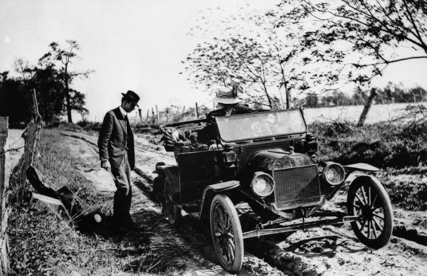 A man fixes a flat tire on an early model Ford automobile. Another man stands to the side of the road, and a woman wearing a hat with a large ribbon waits in the car. Suitcases are piled on the side of the road.