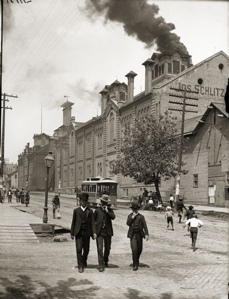 Exterior of Schlitz Brewing Company with two men and a boy in suits and hats in the foreground.