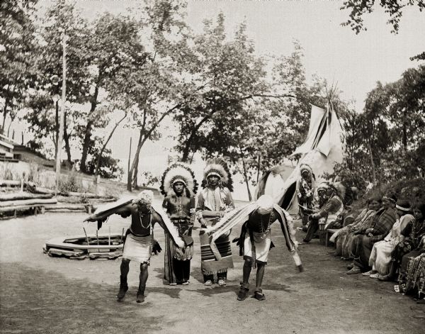 Ho-Chunk Eagle Dance. Two Ho-Chunk men dance in front of two drummers. A group of Ho-Chunk sit on a bench on the right. Tepee in the background.