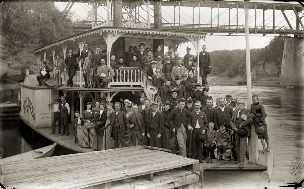 Passengers posing on "Dell Queen" steamboat. There is a bridge in the background.