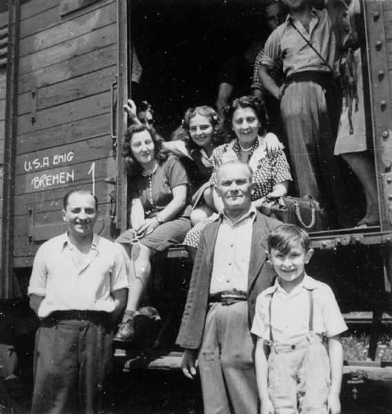 The Weinschenker family poses for a portrait in a train car. From left to right are an unidentified man, an unidentified woman, Frieda Weinschenker (sitting in the doorway of the train car), her mother Klara Weinschenker, her father Chaim Weinschenker (standing in front of Klara) and her brother Milo Weinschenker. They are on their way to Bremerhaven, Germany, to sail to the United States.
