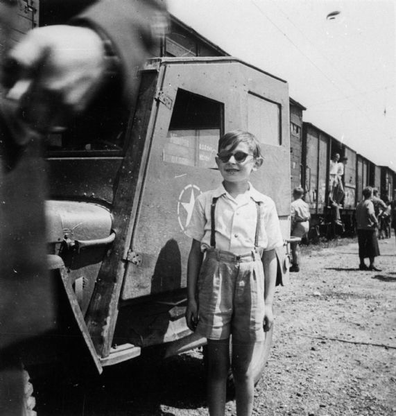 Portrait of a refugee child, Milo Weinschenker, wearing sunglasses. He is standing next to a train, as he and his family were about to leave Germany to sail to the United States.