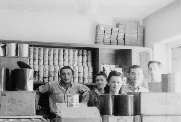Refugees stand  behind supplies in a food warehouse at a displaced persons camp; Germany.

Saul Sorrin was interviewed as part of the Wisconsin Survivors of the Holocaust Interviews project. Sorrin, born in New York in 1919, applied in 1940 for a position with the United Nations Relief and Rehabilitation Administration (UNRRA). He worked with Holocaust survivors as a supply officer for UNRAA team 560 at the Displaced Persons camp Neu Freimann Siedlung in Germany and later, at General Dwight D. Eisenhower's recommendation, Sorrin became the Area Director of the International Refugee Organization based at the Wolfratshausen DP camp in Bad Kissingen.

When asked about this image, Sorrin revealed, "This is the food. They portioned in the food warehouse in one of the camps, food distribution. Now food was cooked, wherever possible, in the individual family units if there was a family. For those who did not have a family we had communal kitchens and these [biksn] or cans, [büksn] of food were distributed as part of a regular worked-out ration. We had dieticians who planned the food distribution according to the UN calorie counts. Each person came with a different colored card which described the number of calories he was entitled to based on whether he was a hard worker or a not so hard worker or a child or whatever. The food came from the German economy mostly and it was supplemented by the United Nations with foods purchased elsewhere. Canned fish for example would come from Sweden, Norway."

Interview by Jean Loeb Lettofsky and David Mandel, March 3, 1980.