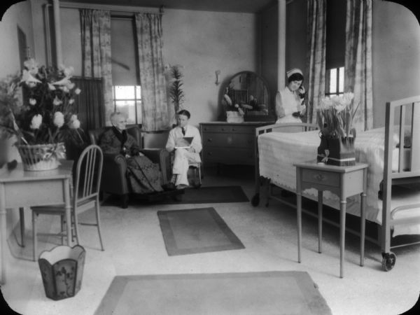 Posed view of a patient room in the recently constructed Methodist Hospital, showing a nurse on the telephone near a bed, and a doctor and elderly patient sitting next to each other.