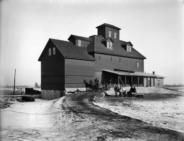 Landscape photograph of the Fairchild grain elevator on a winter's day. There are workers steering two horse-drawn carts in the foreground.
