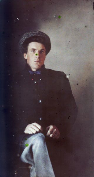 Autochrome portrait of a young man seated with his hands on his knee.  He wears a cap and bow tie.
