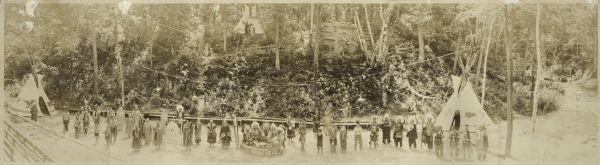 Panoramic view of a large group of Native Americans posed in front of Stand Rock between two tipis.