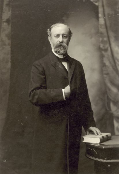 Studio portrait in front of a painted backdrop of William F. Vilas posing with one hand on a law book and the other inside his long overcoat.