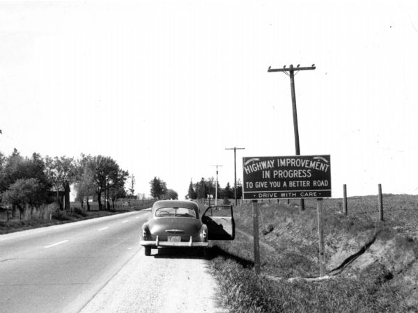 Car stopped on the side of a road with its passenger side door open, next to a sign reading "Highway improvement in progress to give you a better road. Drive with care".