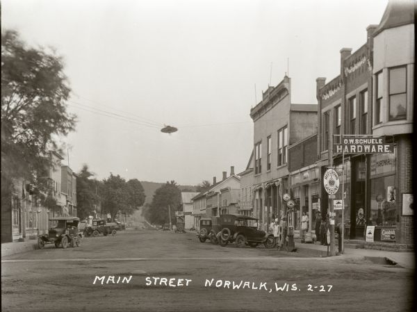 Main Street with several cars parked along the curb. A curbside Red Crown gas pump is visible at the right near the O.W. Schuele Hardware store, and people are standing in the doorways of other storefronts nearby.