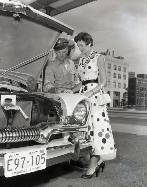 Woman in polka dot dress is looking on as a service station worker checks the oil in her car.