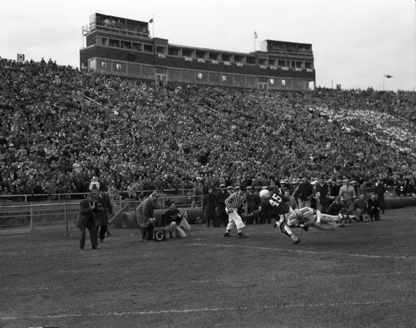 University of Wisconsin-Madison's Danny Lewis narrowly escapes a tackler to score a touchdown vs. Ohio State in a game at Camp Randall Stadium. A large crowd is in the stands, as are photographers and reporters along the sidelines. Wisconsin lost 16 to 13.