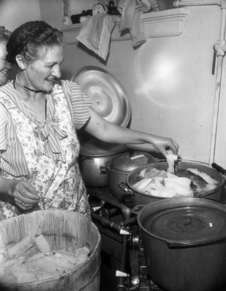Woman wearing an apron in an industrial kitchen and preparing a large pot of lutefisk possibly for a Syttende Mai celebration.