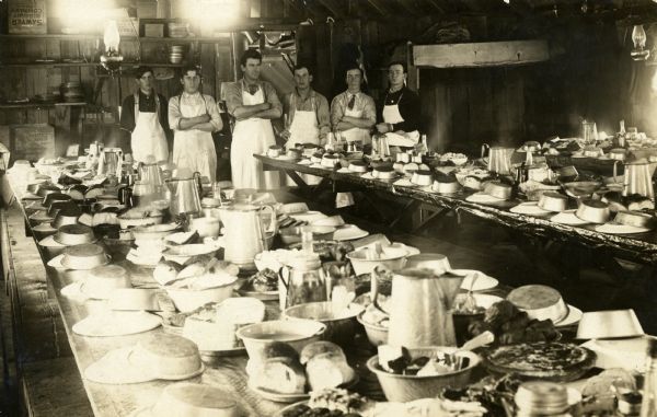 Six cooks wearing aprons stand in a lumber camp dining room with the tables set to serve the crew.