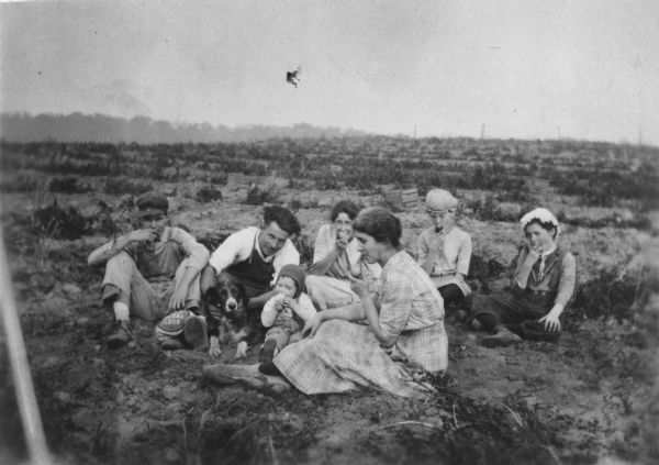 Group of seven people seated in a field and eating raw potatoes. The family dog is also present.