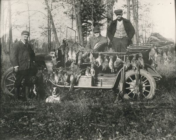 Hunting party of three men with two dogs, guns leaning against a car, and game strung out on the vehicle.