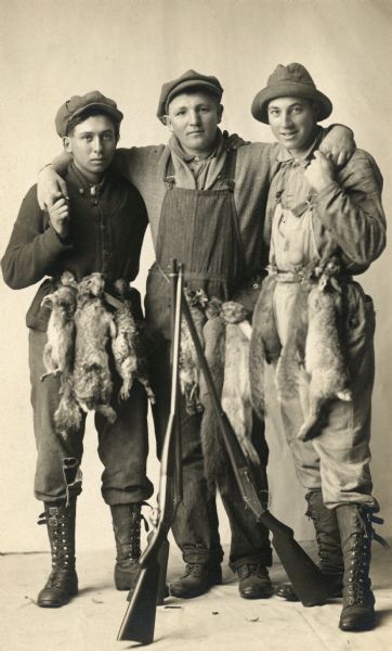Full-length studio portrait of Otto Radke, Andrew Echstein, and Jake Lange posed in front of a painted backdrop. They have their guns leaning in front of them, and squirrels, rabbits, and grouse tied to their belts.