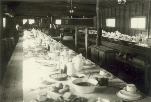 Interior view of the dining room at the Greenwood lumber camp. The long tables are set with dishes and food, and there is a wood stove between the tables.