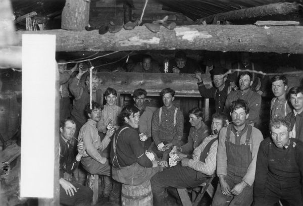 A large group of men, probably logging workers, are shown playing cards in a sleeping shanty.