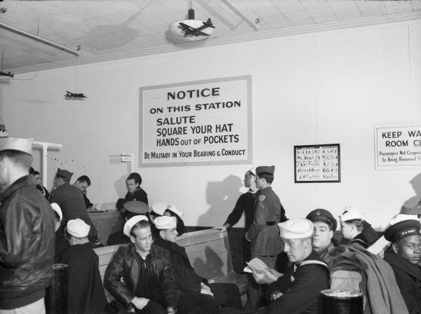 Military personnel in the Naval Air Station waiting room. A sign on the wall reads: "Notice/ On this station salute/ square your hat/ hands out of pockets/ Be military in your bearing and conduct".