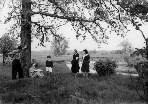 Two men, a young boy, and two women relax by a tree near a stream.