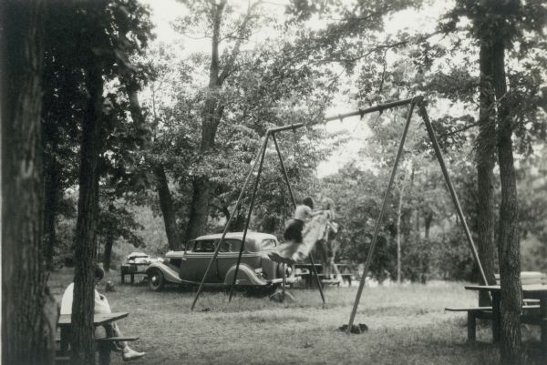 Children playing on swing set in front of an automobile. A man is seated on a picnic table nearby.