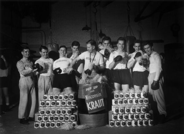 University of Wisconsin freshman boxing team dressed in boxing uniforms and gloves standing behind stacked cans of sauerkraut with a sign that reads "Pep Up with Wisconsin Kraut".
From left to right: Jim Walsh, Art Walsh, Omar Crocker, Truman Torgerson, John Walsh, Nick Lee, Ray Kramer, Woody Swancutt, and Gene Rankin.