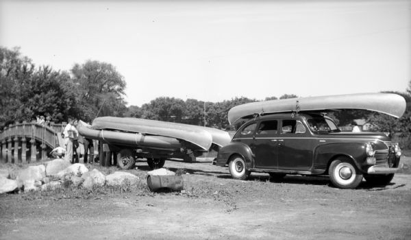 Group of people standing near a car with a trailer carrying canoes at Vilas Park. There are people standing on a bridge over water in the background.