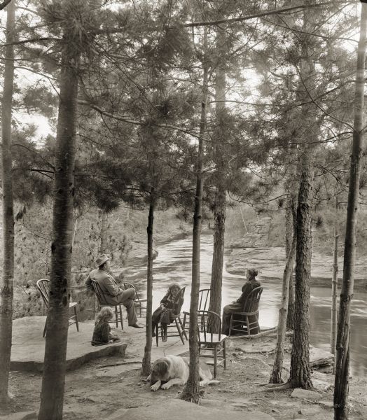 Crandall family, including a dog, sitting under trees on a cliff overlooking a river. From left to right: George Crandall, Lois Crandall, Phyllis Crandall, Nellie Bennett (Crandall).