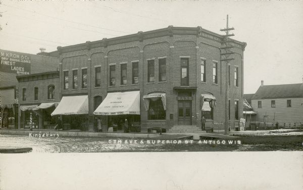 Exterior view across intersection toward Ed. E. Williams' Drug Store, Langlade National Bank, and The Gismonda Soda Fountain at 5th Avenue and Superior Street. A man is standing at the corner on the steps of the bank. Caption reads: "5th Ave. & Superior St., Antigo, Wis."