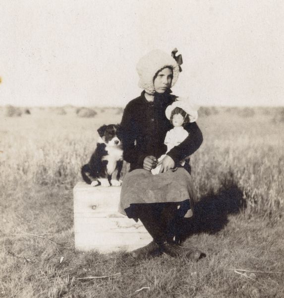 Miss Irene Baudle cradles her doll while sitting next to her puppy in front of the family wheat field.