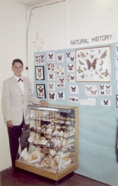 A young man poses in front of a Natural History exhibit which includes a glass case of shells, butterflies and moths mounted on the wall. He donated his collection to Upper Iowa University.