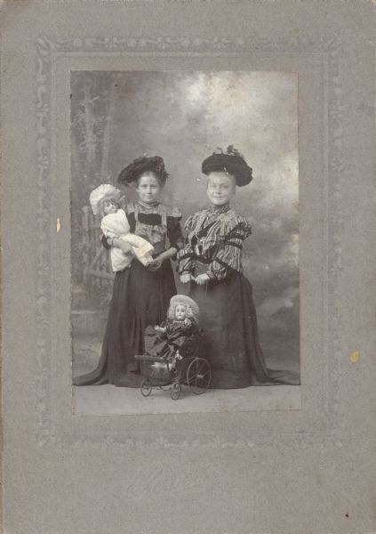 Two young girls dressed up as women in front of a photographer's backdrop. The girl on the left holds a doll while the girl on the right pushes her doll in a stroller.