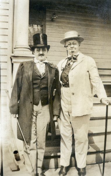 Two women dressed up in men's attire, including top hats, eyeglasses, fake moustache, and canes, stand on the steps of porch of a private residence.